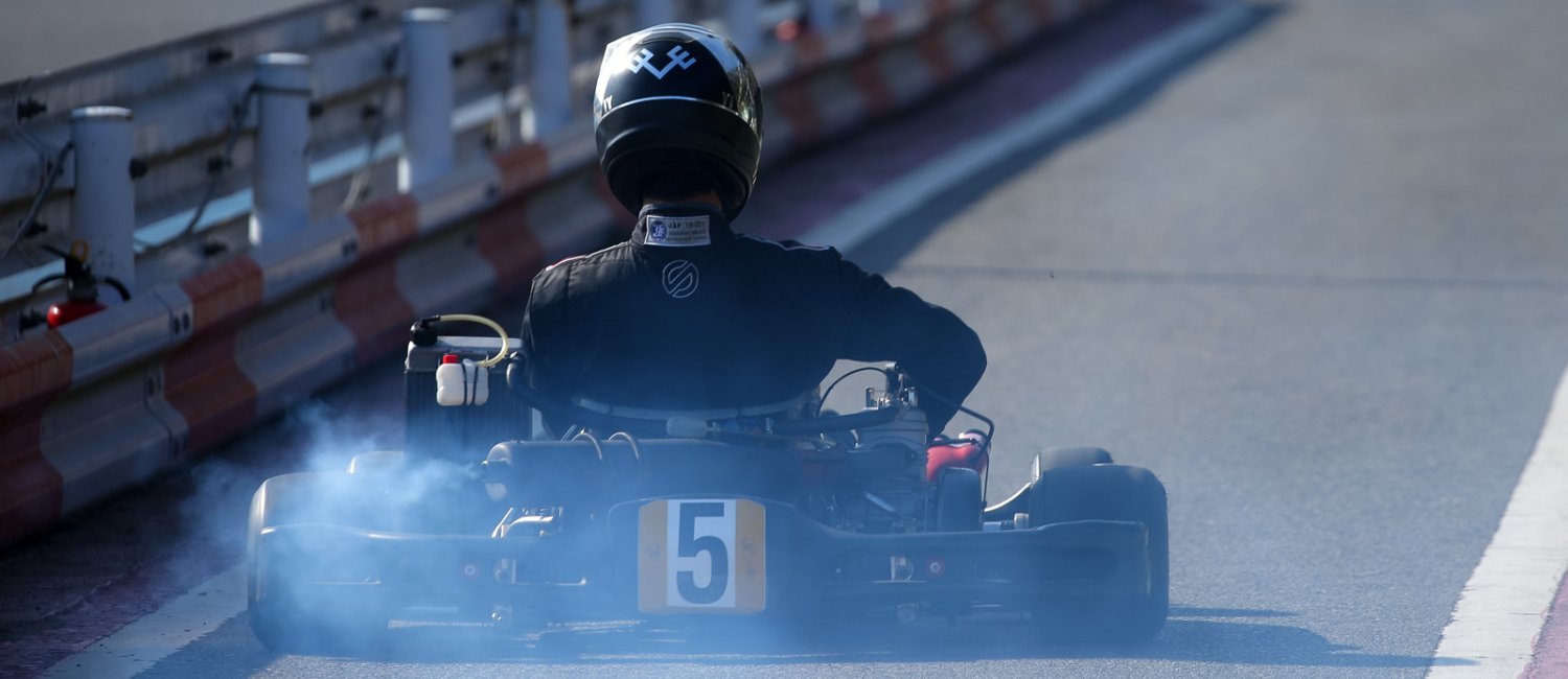 A view from behind a go-kart race driver in a black racing suit. The go-kart blows out a thick, blue cloud of exhaust gas.