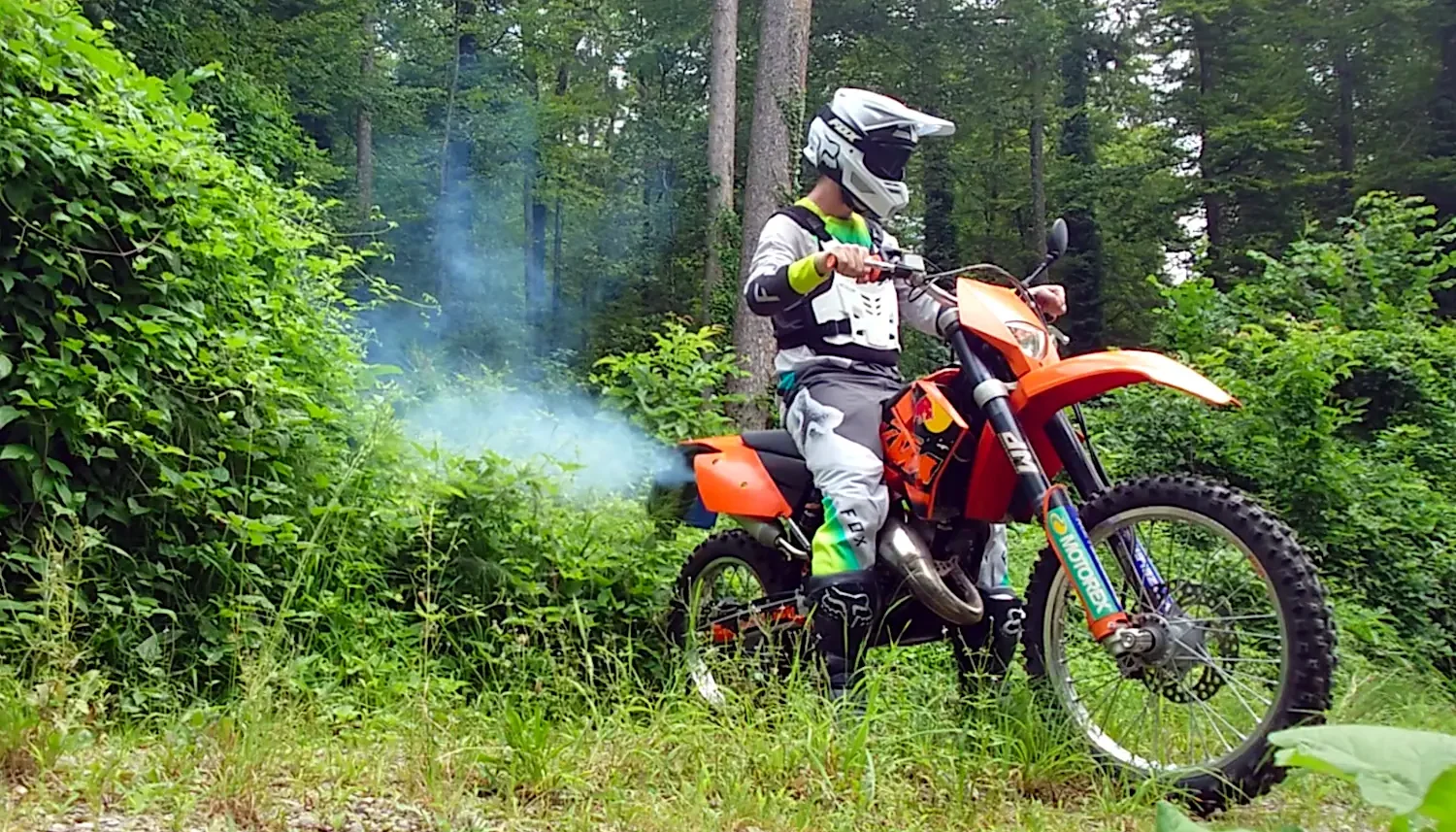A young motocross rider is revving a two-stroke dirtbike, blowing thick, blue clouds of exhaust fumes into a pristine, lush forest.