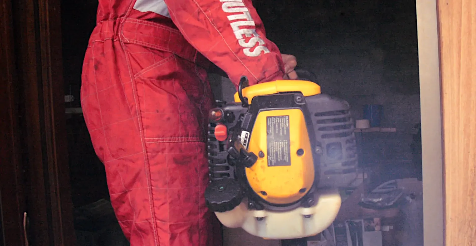 A young man holding a yellow two-stroke string trimmer. The exhaust outlet is pointed at the camera, with a faint blue haze pouring out of it.