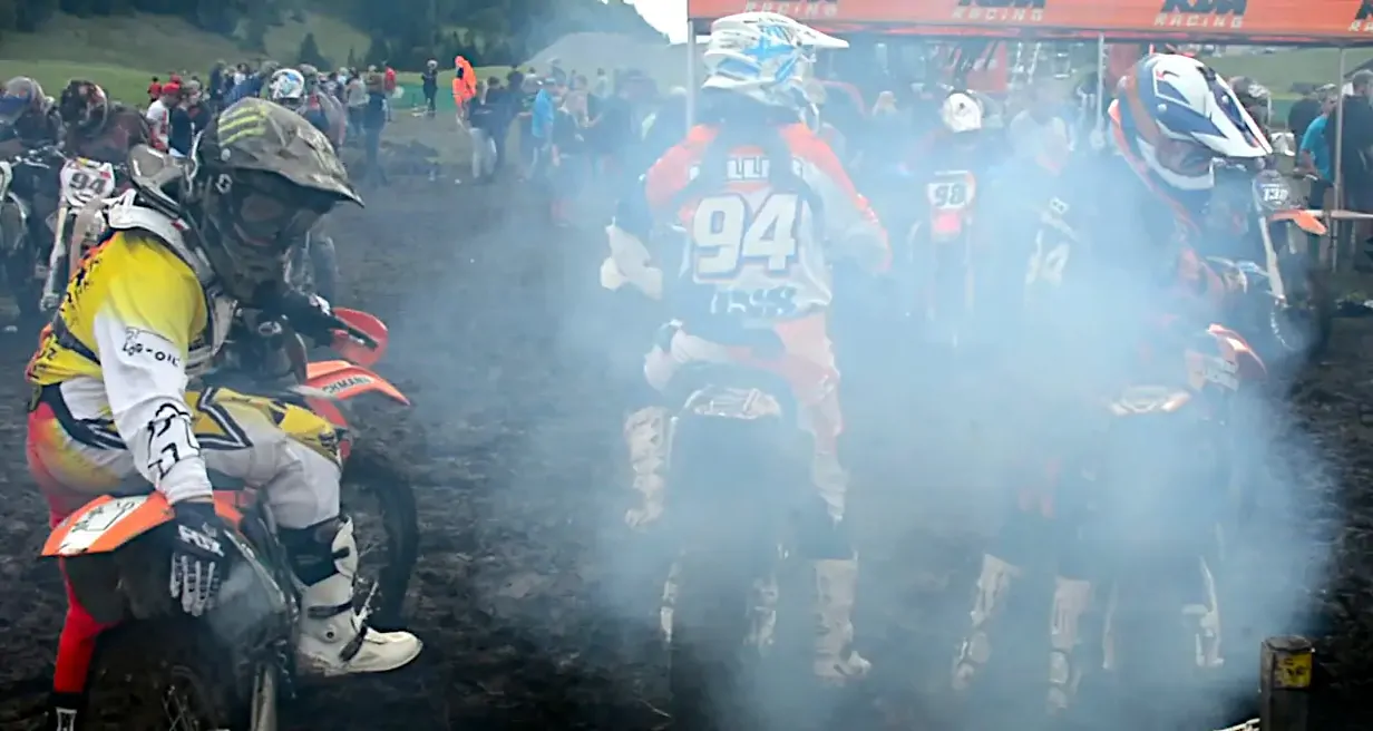 A group of motocross riders on their dirtbikes from behind, with massive amounts of thick, blue exhaust fumes obscuring most of the view.
