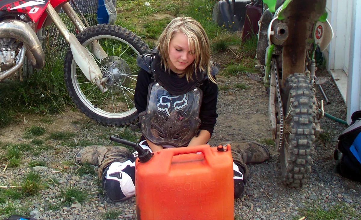 A young blonde girl wearing motocross pants, boots, and chest protector, next to two dirtbikes and a large, orange gas can.
